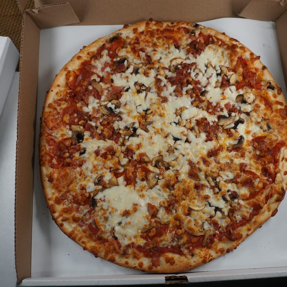 Large freshly baked pizza topped with melted cheese, pepperoni, bacon, mushrooms, and red pepper flakes in an open cardboard box.
